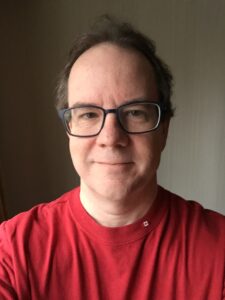 A picture of Doug taken in 2019. He is facing the camera with a small smile on his face. He is white, wearing large glasses with squarish lenses and a red t-shirt. He has a tiny Canadian flag pin on his collar.