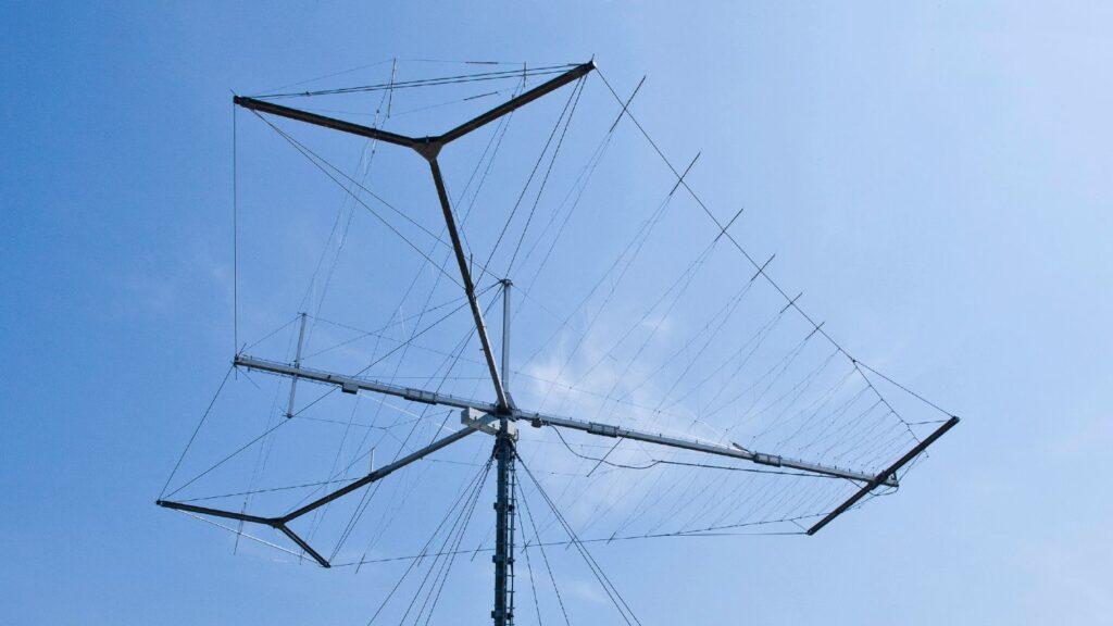 Large log-periodic HF antenna mounted on a tall mast, its triangular support arms and multiple parallel wire elements stretched out against a clear blue sky.
