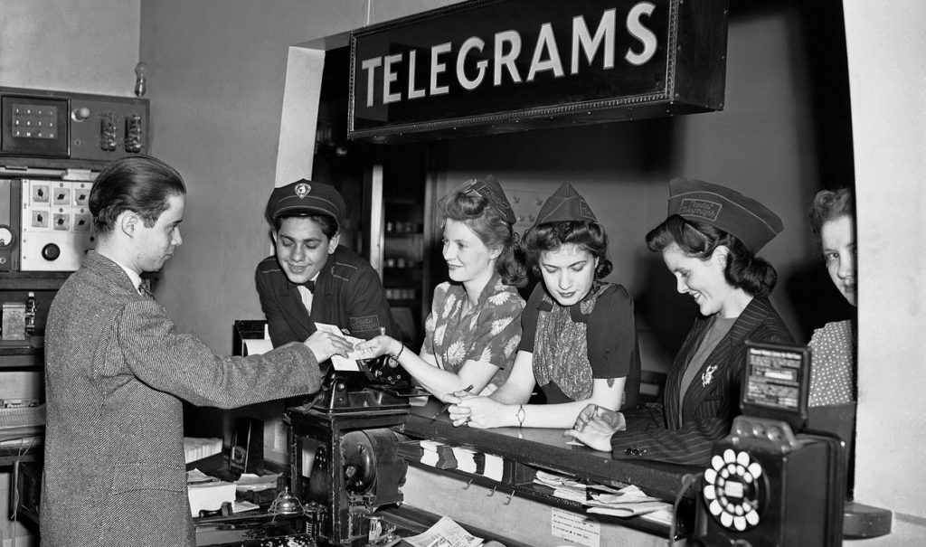 A 1942 black-and-white Associated Press photograph showing a young man handing a message across a counter at a Western Union telegram office. Behind the counter, four women and one male employee—dressed in wartime-era uniforms and patterned dresses—process telegrams and interact with customers. A large “TELEGRAMS” sign hangs overhead, and vintage communication equipment and a rotary telephone frame the busy, high-energy scene typical of World War II-era message centers.